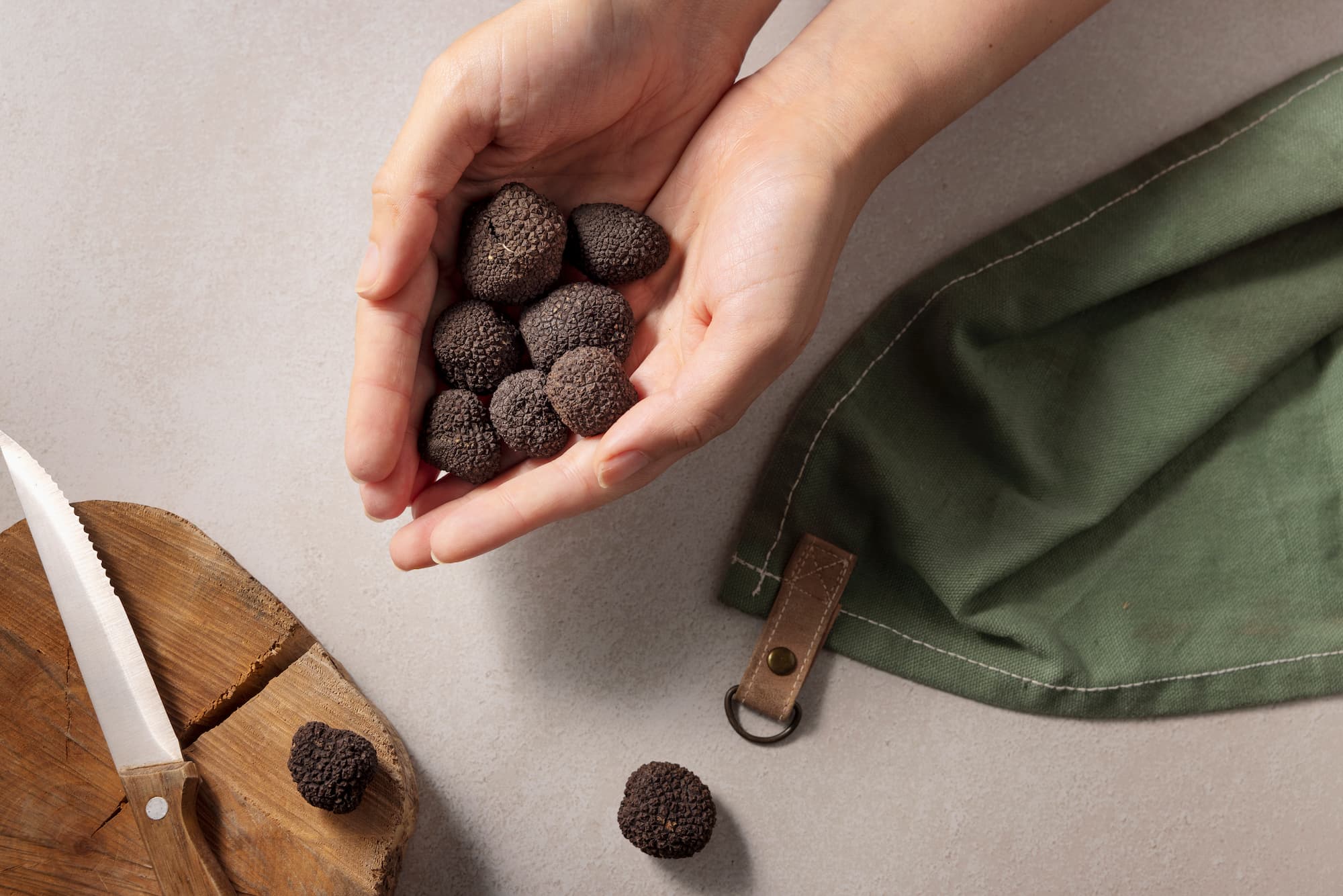 deux mains tenant sept têtes de truffes au dessus d'une table avec un couteau sur une planche à découper en bois, deux autres truffes sur la table et un torchon en tissus vert